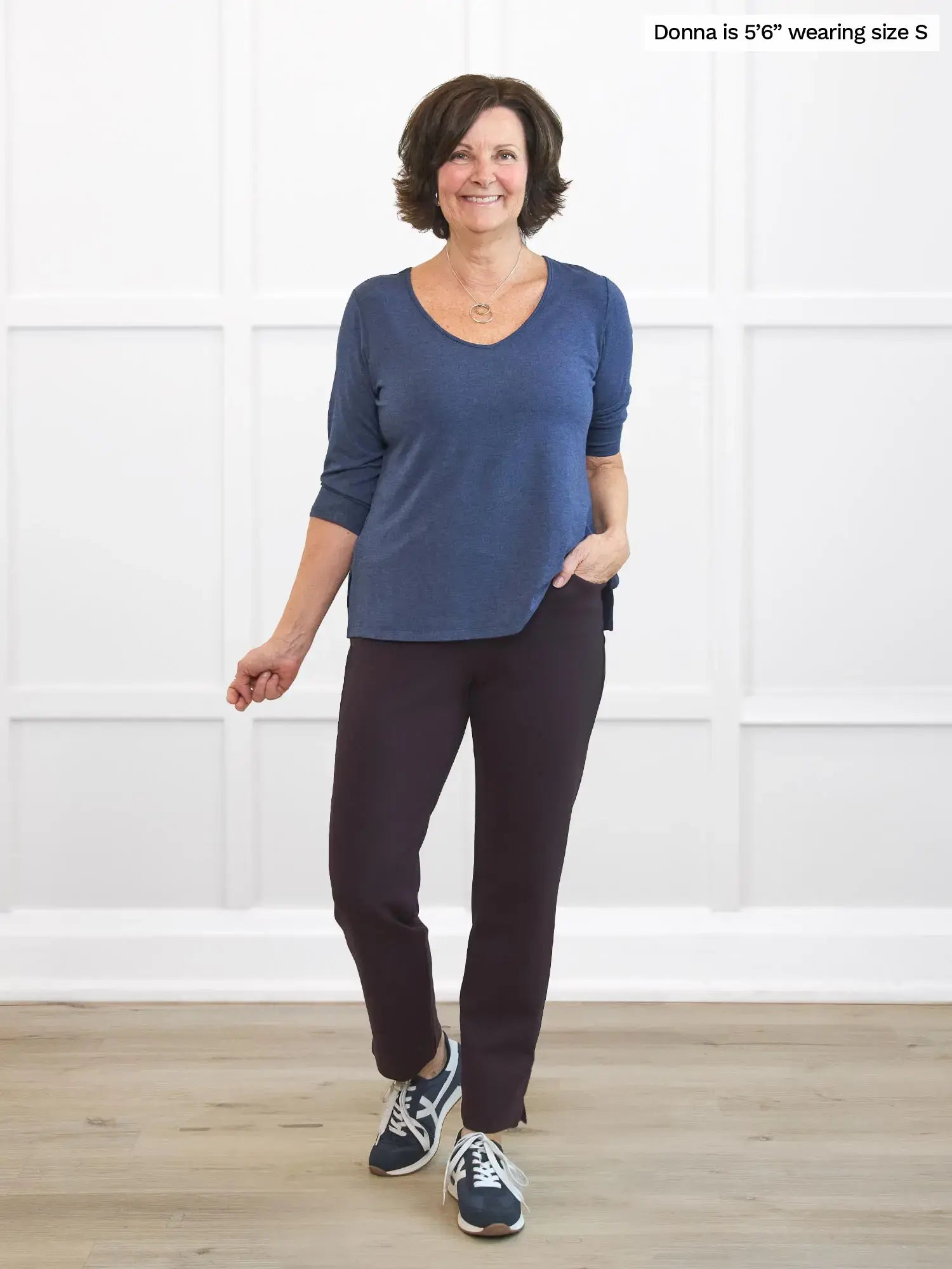 Woman wearing a blue top and dark brown pants standing against a white wall.