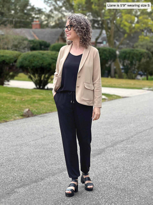 Miik model Liane (5'9", small) smiling and looking away wearing Miik's Perle open-back sleeveless jumpsuit in black with a machine washable blazer in wheat colour 