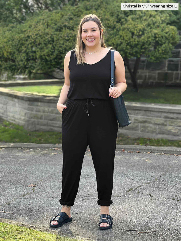 Miik model Christal (5'3", large) smiling wearing Miik's Perle open-back sleeveless jumpsuit in black with a tote bag an sandals 