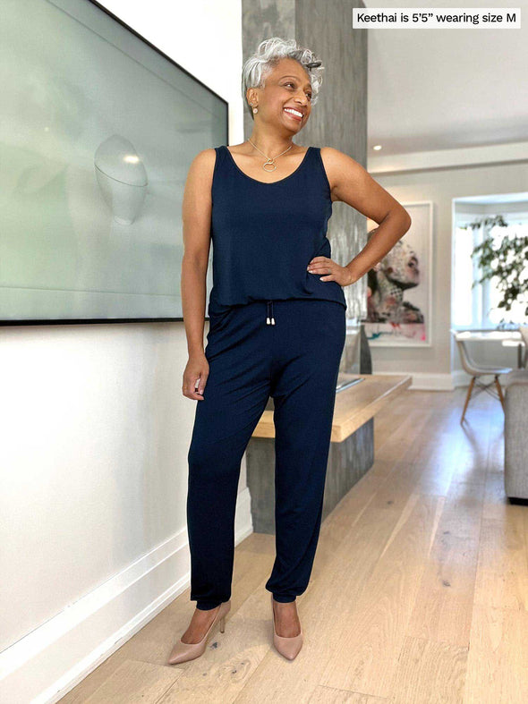 Woman standing in a living room looking away wearing Miik's Perle open-back sleeveless jumpsuit in navy