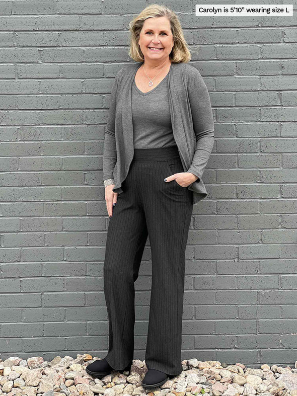 Woman standing in front of a wall wearing Miik's Reed high waisted pant with pockets in charcoal pinstripe with a grey top and a matching cardigan.