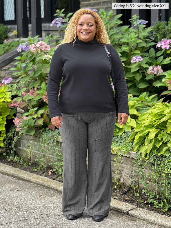 Miik model Carley (five feet two, xxlarge) smiling in front of a garden wearing Miik's Reed high waisted pant with pockets in granite melange pinstripe along with a graphite long sleeve top