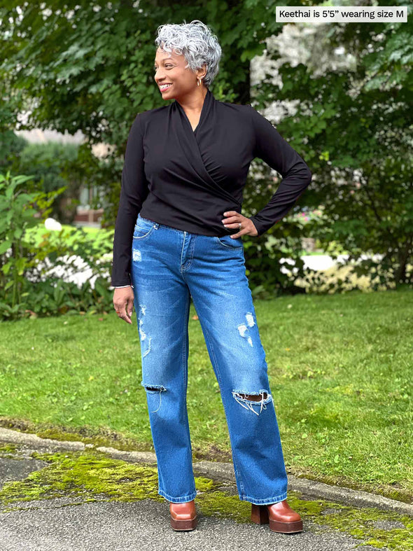 Miik model Keethai (five feet five, medium) smiling and looking away wearing Miik's Reni ruched faux-wrap blouse in black with jeans 