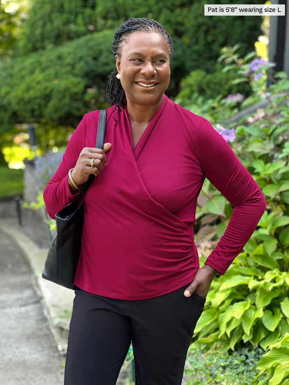 Miik model Pat (five feet eight, large) smiling while standing in front of a garden wearing Miik's Reni ruched faux-wrap blouse in bordeaux along with a black pant