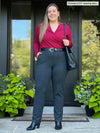 Miik model Christal (five feet three, large) smiling in front of a door wearing Miik's Reni ruched faux-wrap blouse in bordeaux with a charcoal pant, black belt, purse and boots