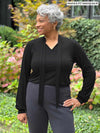 Miik model Keethai (five feet five, medium) standing in front of a garden looking away wearing Miik's Rhoda tie neck blouse black untied with a graphite flare pant
