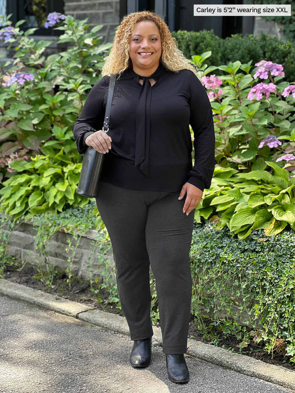 Miik model Carley (five feet two, xxlarge) smiling wearing Miik's Rhoda tie neck blouse in black along with a charcoal pant, black boots and purse 
