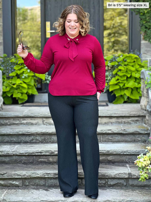 Miik model Bri (five feet five, xlarge) standing in front a doorway looking down wearing Miik's Rhoda tie neck blouse in bordeaux with a charcoal pinstripe pant and sunglasses