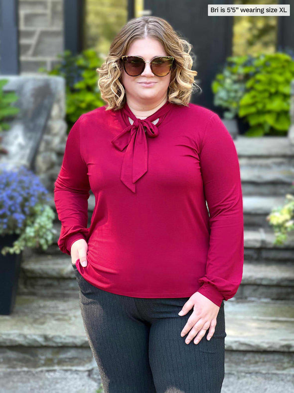 Miik model Bri (five feet five, xlarge) standing in front a doorway wearing Miik's Rhoda tie neck blouse in bordeaux with a charcoal pinstripe pant and sunglasses 