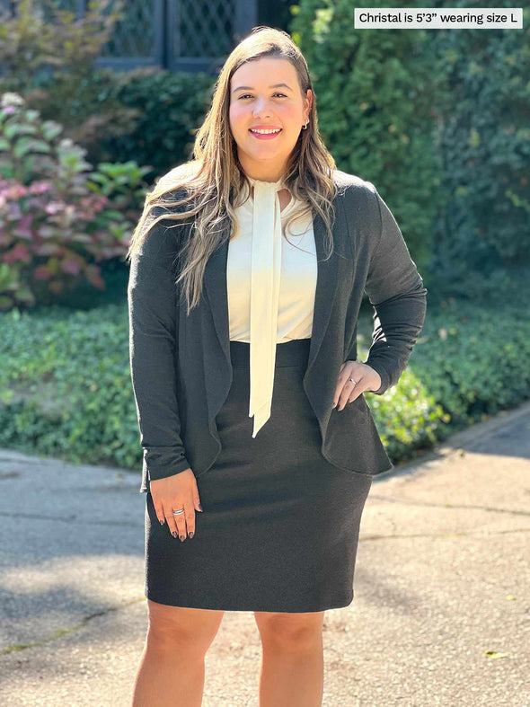 Miik model Christal (5'3", large) smiling wearing Miik's Rhoda tie neck blouse in natural along with a pencil skirt and a soft blazer in charcoal 