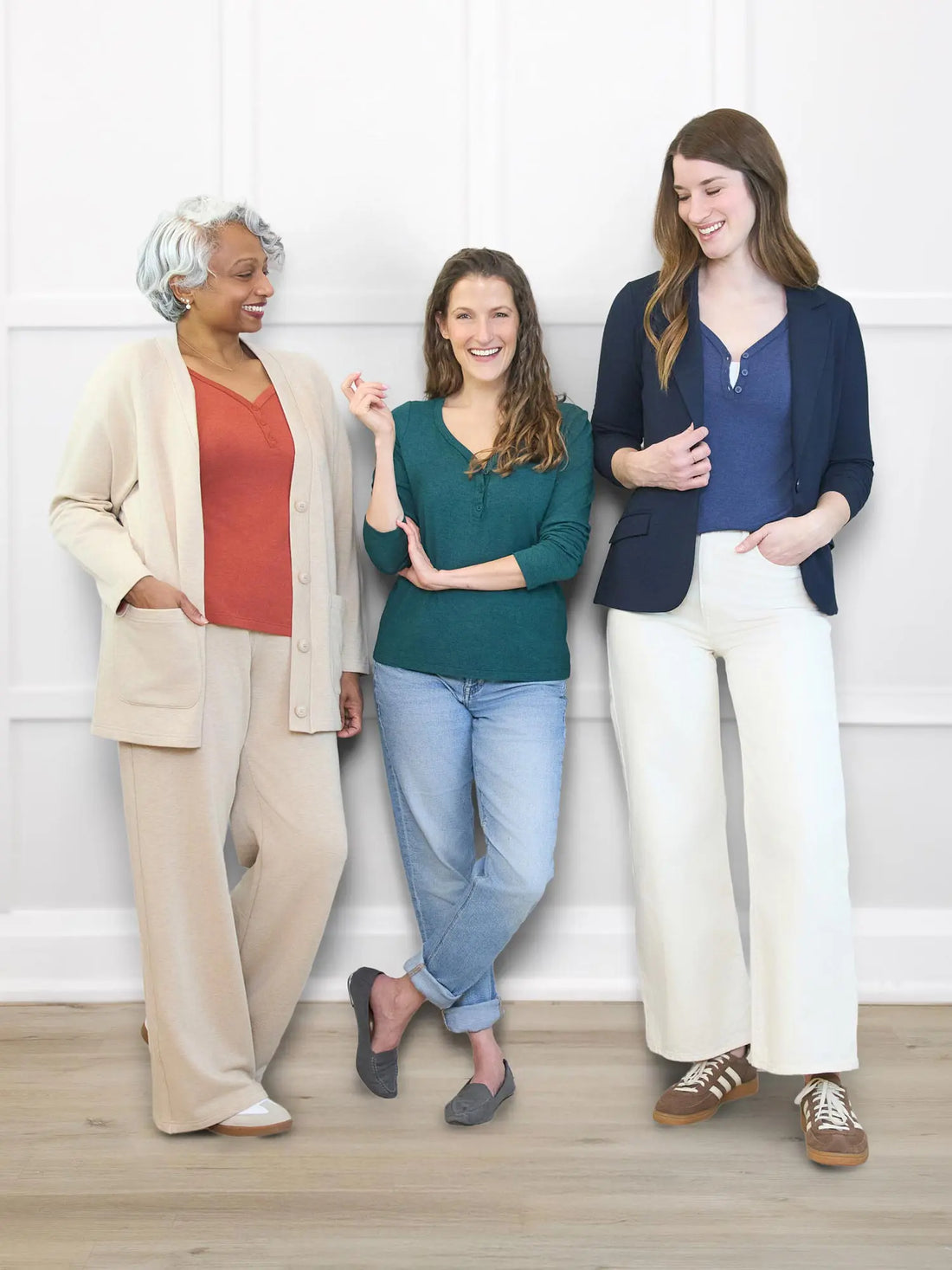 Three women standing together against a white wall, wearing casual clothing.