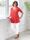 Miik founder Donna (five feet six, size small) smiling while standing next to a window wearing a white jeans along with Miik's Rocelle half-sleeve scoop neck tunic in papaya wide pinstripe and brown sandals