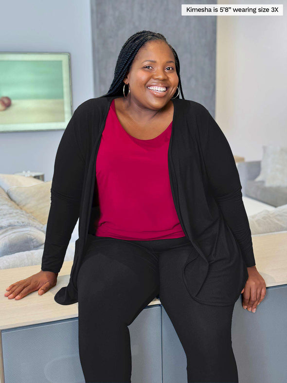 Miik model plus size Kimesha (5'8", size 3x) smiling and sitting on top of a living room furniture wearing Miik's Rory waterfall cardigan in black with a pant in the sae colour with a tank top in bordeaux