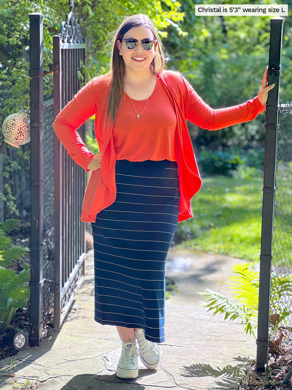 Miik model Christal (five feet three, large) standing next to a backyard gate smiling wearing a pinstripe navy midi skirt along with Miik's Rory waterfall cardigan in papaya melange with a tank top in the same colour
