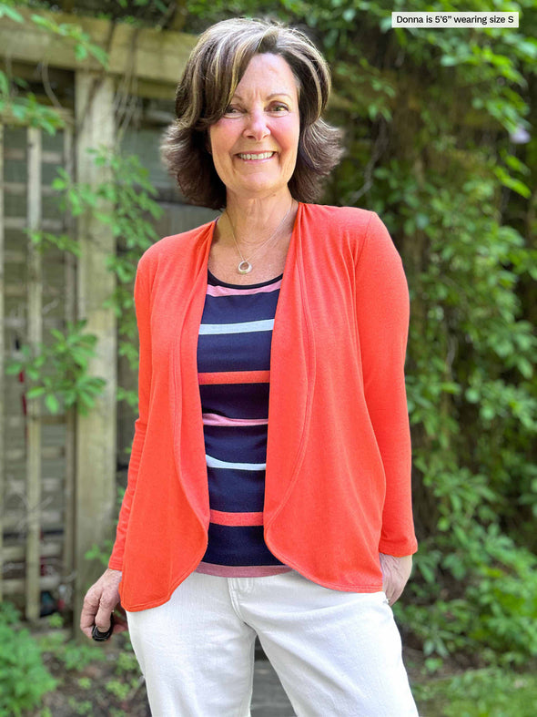 Woman standing in nature wearing Miik's Rory waterfall cardigan in orange over a striped top and white pants.