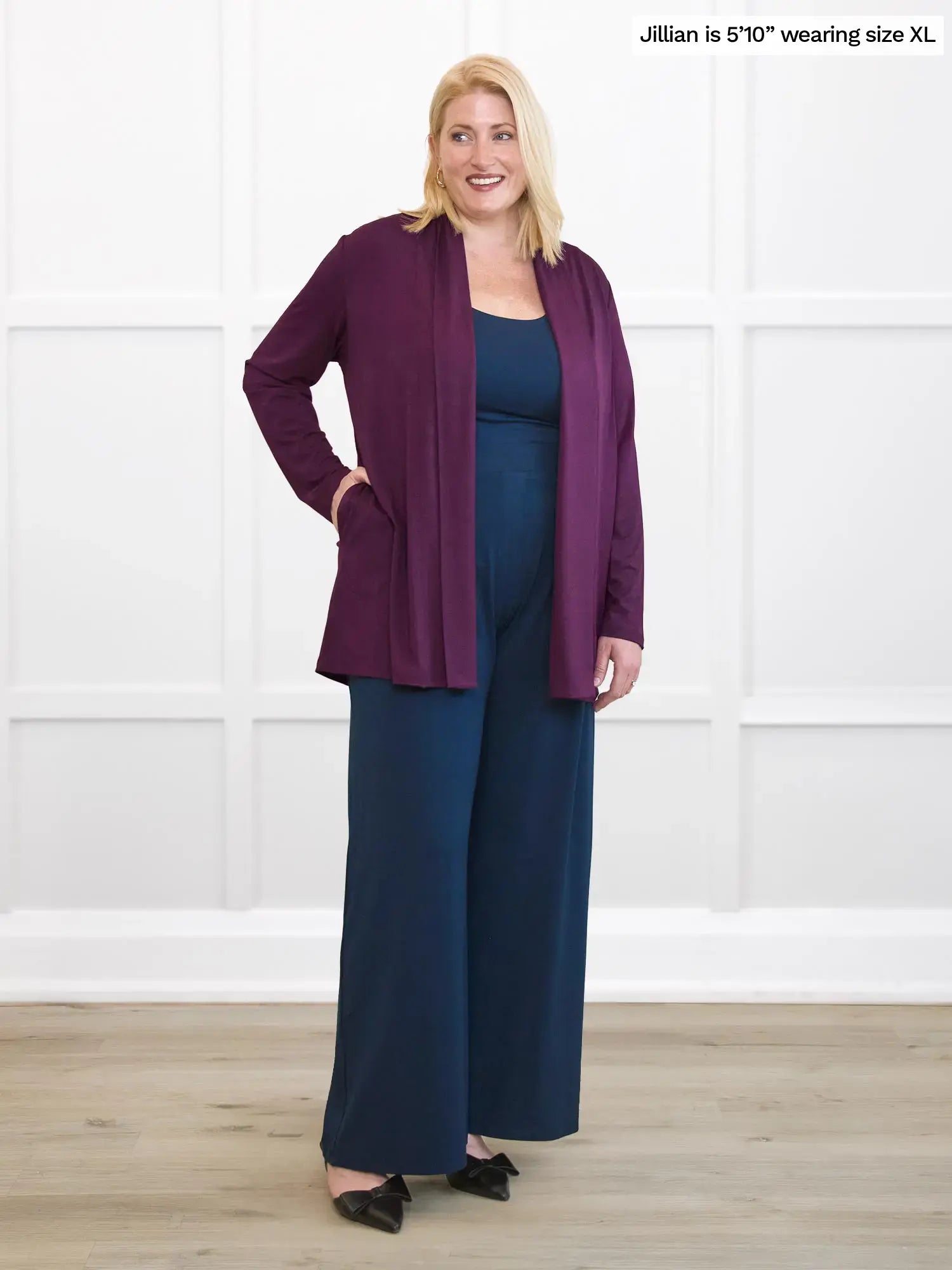 Woman wearing a purple cardigan and navy pants against a white background 