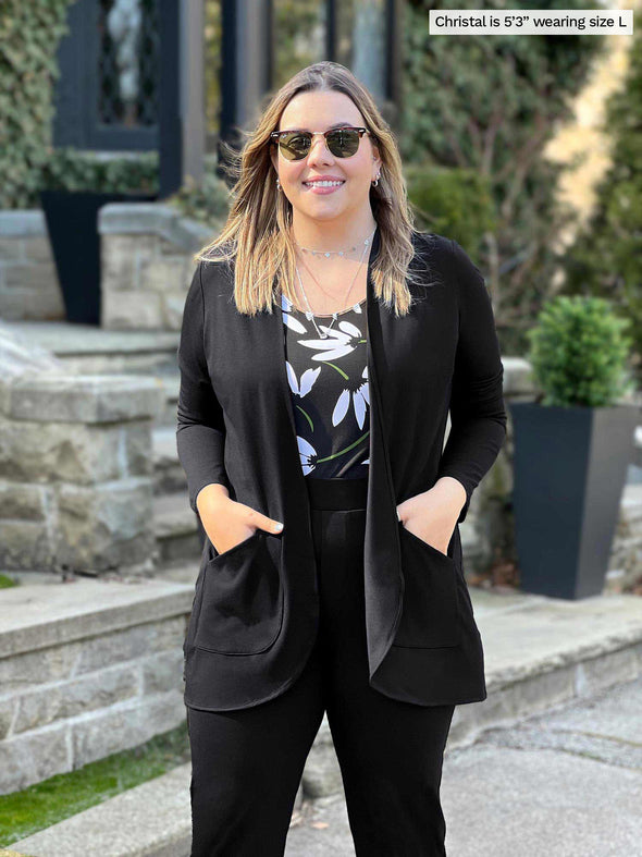 Miik model Christal (5'3", large) smiling wearing Miik's Sade open-front pocket cardigan in black with a pant in the same colour and a printed tank  and sunglasses 