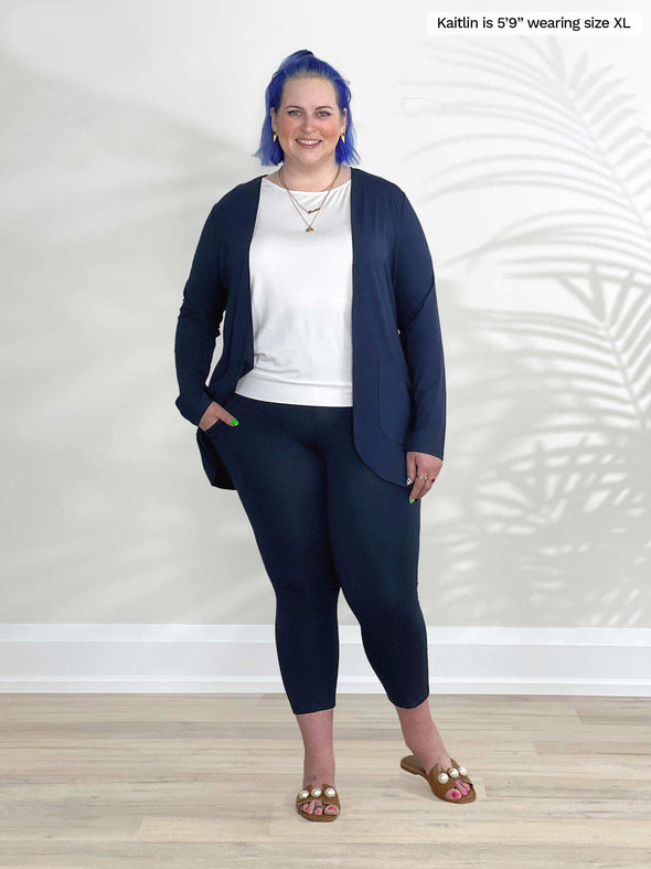 Miik model Kaitlin (5'9", xlarge) smiling and looking away while standing in front of a white wall wearing Miik's Sade open-front pocket cardigan in navy with a capri pocket legging in the same colour and a white tee