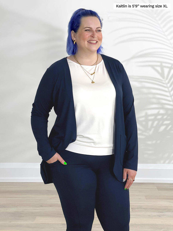 Miik model Kaitlin (5'9", xlarge) smiling and looking away while standing in front of a white wall wearing Miik's Sade open-front pocket cardigan in navy with a capri pocket legging in the same colour and a white tee 