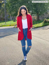 Miik model Yasmine (5'0", xsmall, petite) smiling while looking down wearing Miik's Sade open-front pocket cardigan in poppy red, a white tank and ripped jeans 
