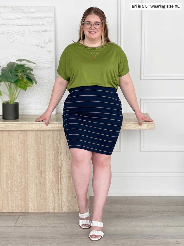 Miik model Bri (5'5", xlarge) smiling while leaning against to a table wearing a green moss shirt with Miik's Salma striped pencil skirt in navy wide pinstripe 