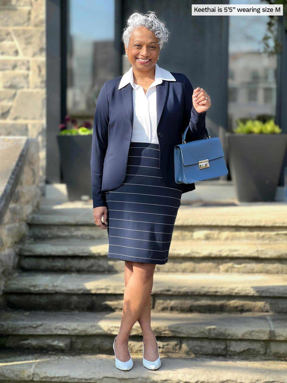 Miik model Keethai (5'5", medium) smiling while standing on a doorway stairs wearing Miik's Salma striped pencil skirt in navy wide pinstripe, a collared shirt in white and a blazer in navy