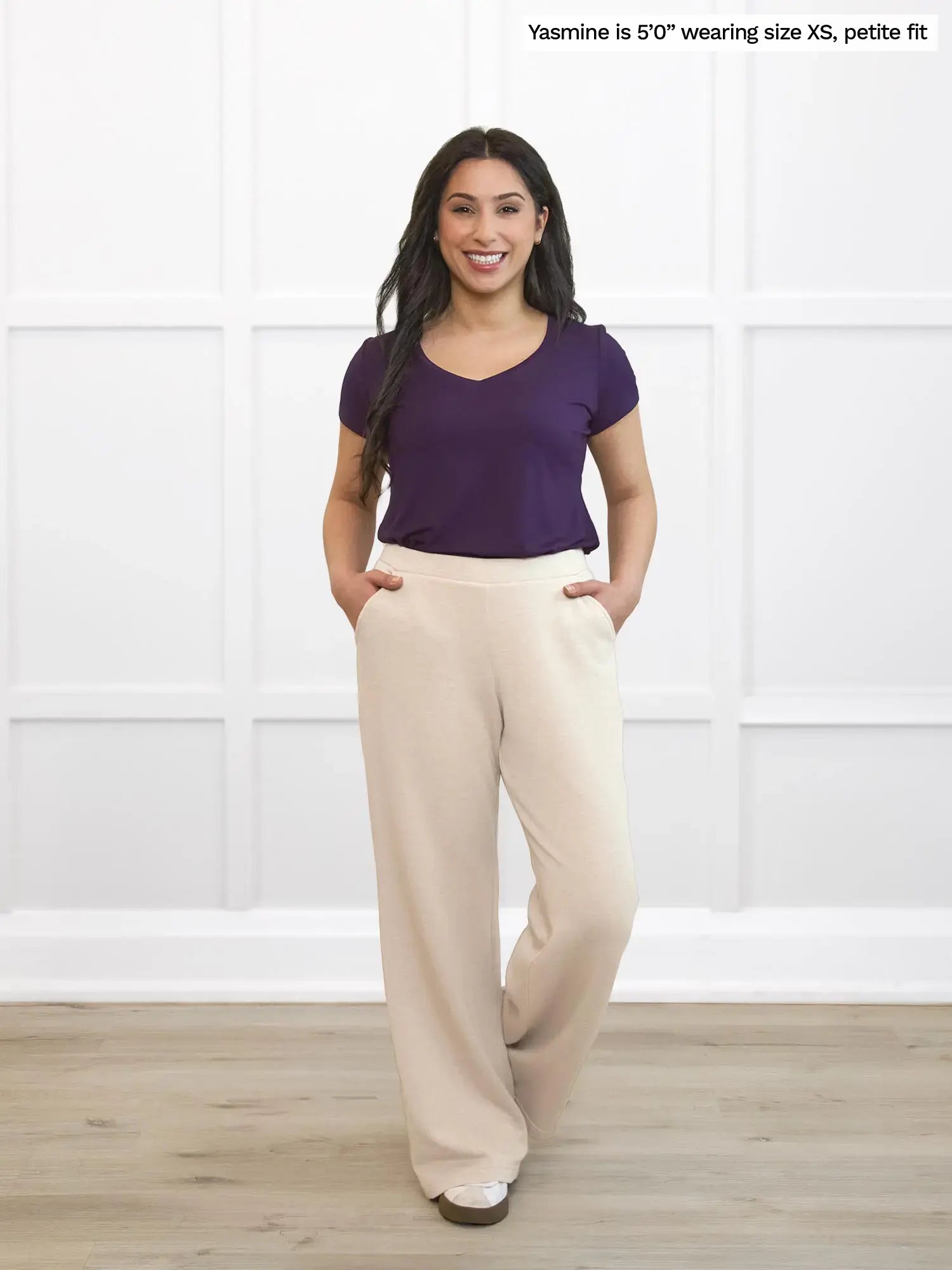 Woman wearing a purple top and beige pants standing against a white wall. 