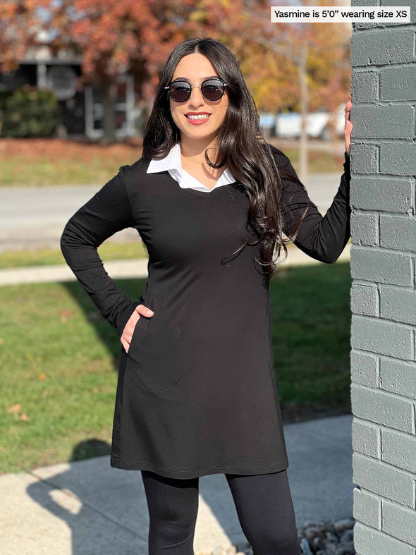 Woman standing next to a building wearing Miik's Saya v-neck pocket tunic in black over a collared shirt with leggings.
