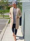 Woman walking next to a wall wearing Miik's Serena long coat in beige with a tunic and leggings.