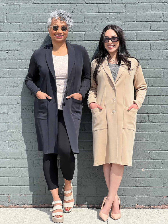 Miik models Keethai and Yasmine standing in front of a brick wall smiling wearing Miik's Serena long coat with pockets in the two colours available: graphite and wheat 