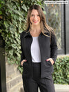 Miik model Johanna (5'6", xsmall) smiling wearing Miik's Shaelyn full zip luxe fleece jacket in black opened along with a white tank and a black dress pant 