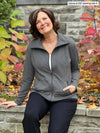 Miik founder Donna (5'6", small) smiling and sitting in front of a garden wearing Miik's Shaelyn full zip luxe fleece jacket in granite in her actual size. She is also wearing it with a black pant and a white tank 