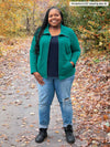Miik model plus size Kimesha (5'8", size 3x) smiling with one hand on pocket wearing Miik's Shaelyn full zip luxe fleece jacket in jade melange with a navy tank and ripped jeans 