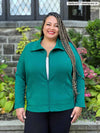 Miik model plus size Sarita (5'7", size 3x) smiling while standing in front of a window/garden wearing Miik's Shaelyn full zip luxe fleece jacket in jade melange with a white tank and navy pants 