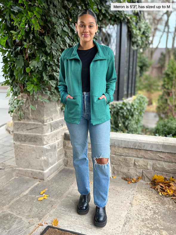 Miik model Meron standing in front of a greener bush wearing a black tank, ripped jeans and Miik's Shaelyn full zip luxe fleece jacket in jade melange. Meron has sized up the jacket to M. She is also wearing it opened