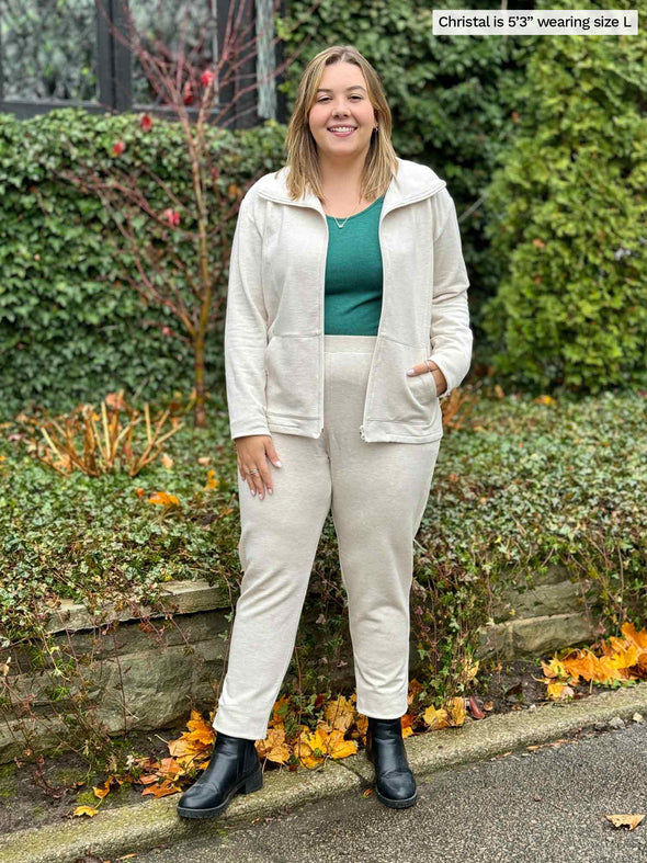 Miik model Christal (5'3", large) smiling while standing in front of a garden wearing Miik's Shaelyn full zip luxe fleece jacket in oatmeal melange, along with a jogger in the same colour and a long sleeve tee in jade melange 