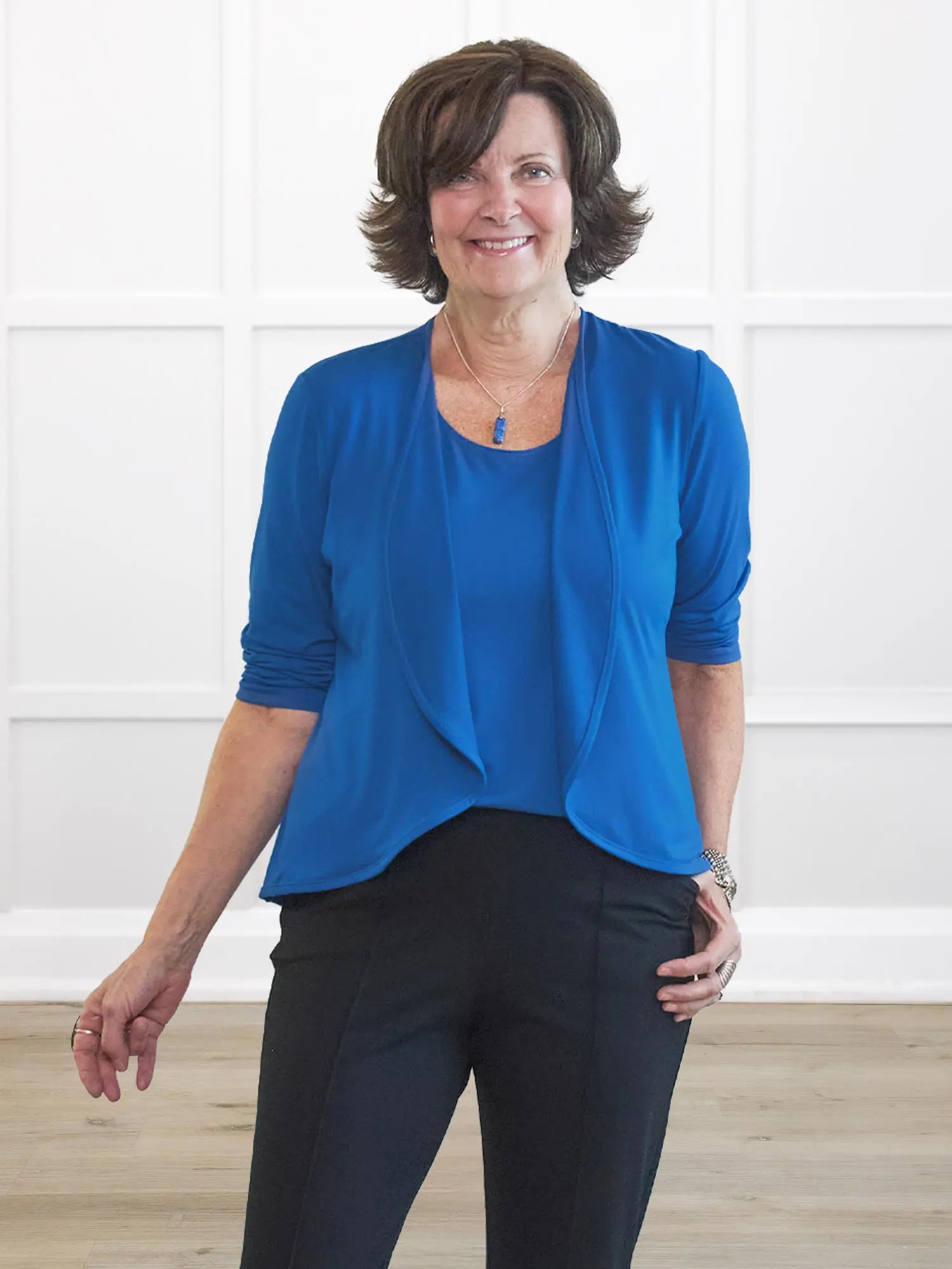 Woman wearing a blue top and black pants against a white wall. 