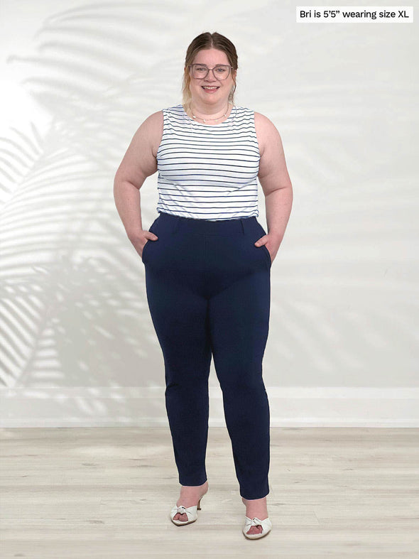 Miik model Bri (5'5", xlarge) smiling while standing in front of a white wall wearing Miik's Shandra reversible tank top - coastal stripe tucked in a navy dress pant