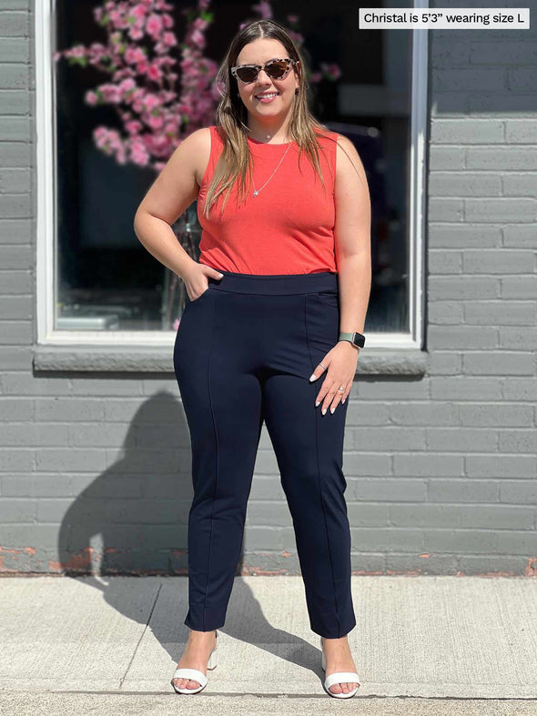 Miik model Christal (five feet three, large) smiling standing in front of a window/brick wall wearing Miik's Shandra reversible tank top in papaya melange with an ankle pant in navt