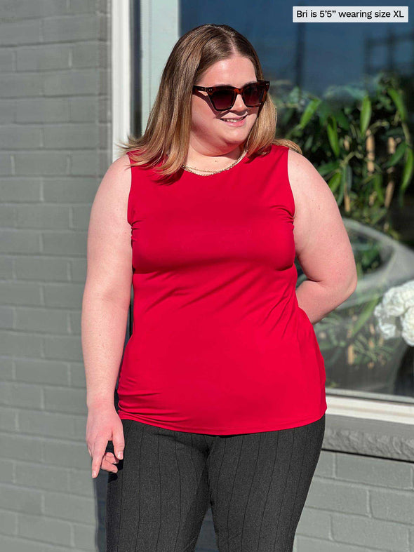 Miik model Bri (5'5", xlarge) smiling while standing in front of a window wearing Miik's Shandra reversible tank top in poppy red along with a pinstripe charcoal pant and sunglasses 