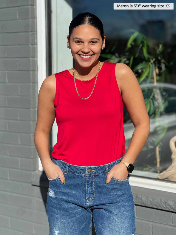 Miik model Meron (5'3", xsmall) smiling while standing in front of a window with both hands on pockets wearing Miik's Shandra reversible tank top in poppy red and jeans