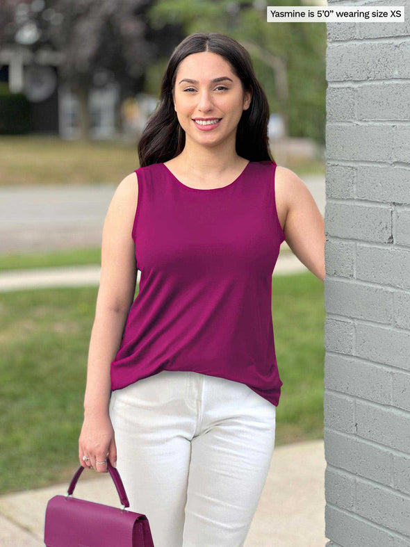 Miik model Yasmine (five feet tall, xsmall, petite) standing next to a brick wall wearing Miik's Shandra reversible tank top in ruby along with a white jeans