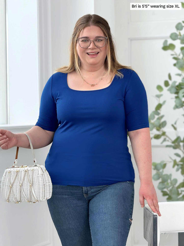 Miik model Bri (5'5", xlarge) smiling wearing Miik's Shani reversible half sleeve square neck top in ink blue with jeans and a white purse 
