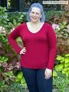Woman standing in front of plants wearing Miik's Shannon long sleeve tee in red with navy leggings