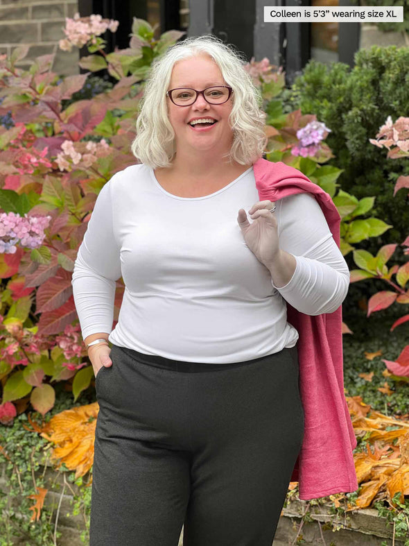 Miik model Colleen (5'3", xlarge) smiling wearing Miik's Shannon long sleeve tee in white with a charcoal fleece jogger and a pink cardigan over her shoulder 