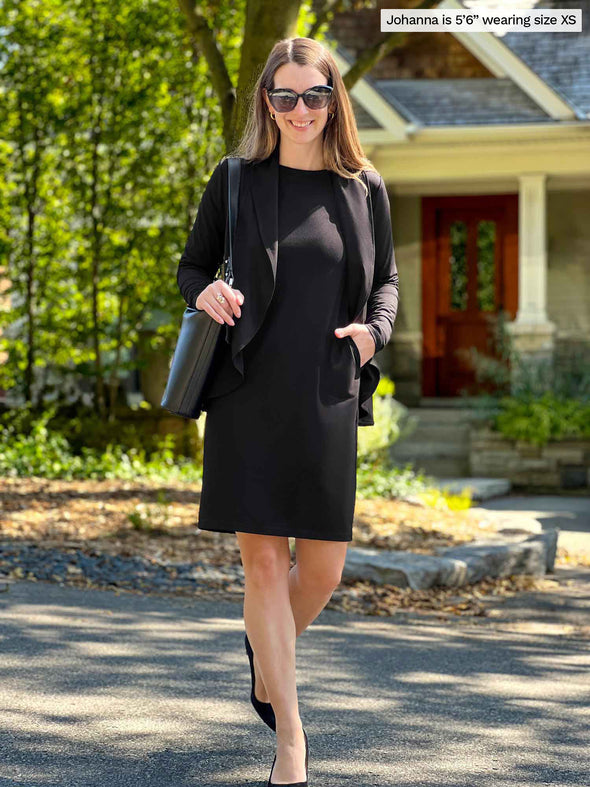 Miik model Johanna (5'6", xsmall) smiling while walking with hand on pocket wearing a black soft blazer cardigan along with Miik's Shena long sleeve boatneck pocket dress in the same colour