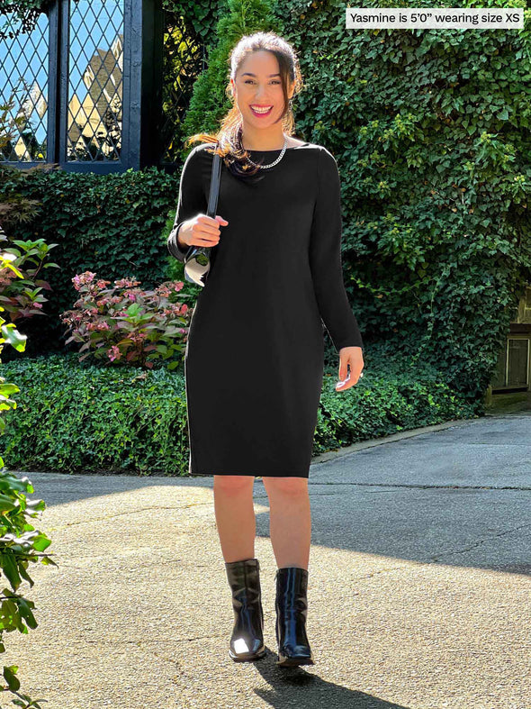 Miik model Yasmine (5'0", xsmall, petite) smiling wearing Miik's Shena long sleeve boatneck pocket dress in black with a shoulder bag and boots in the same colour