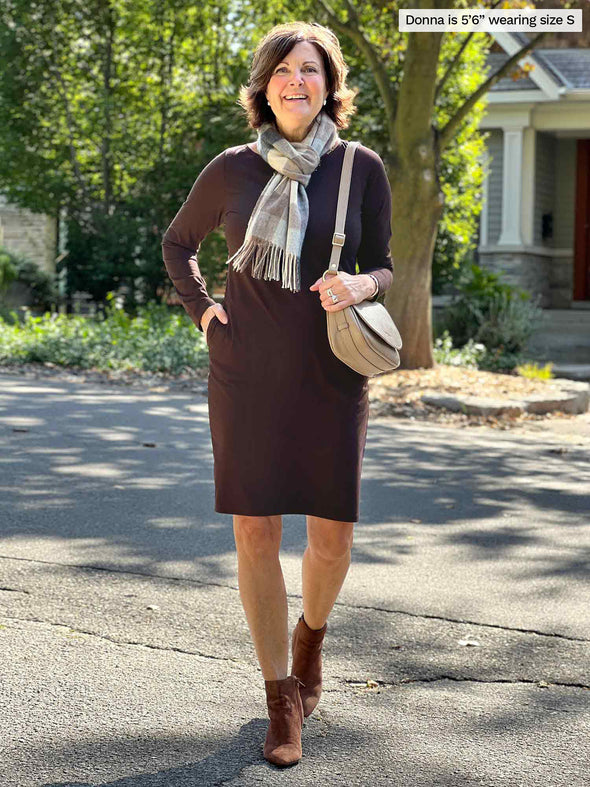 Miik founder Donna (5'5", small) smiling while walking wearing Miik's Shena long sleeve boatneck pocket dress in dark chocolate brown with a scarf with neutral tones, brown boots and purse