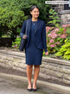 Miik model Meron (5'3", xsmall) looking away while standing in front of garden wearing Miik's Shena long sleeve boatneck pocket dress in navy with a matching colour blazer 
