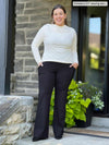 Miik model Christal (five feet three, large) smiling in front of a door wearing Miik's Sierra high waisted pocket pant in black with a long sleeve top in natural
